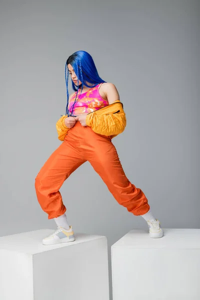 Volle Länge, Mode nach vorne, junge Frau mit blauen Haaren posiert in Pufferjacke und orangefarbener Hose auf grauem Hintergrund, steht auf weißen Würfeln, leuchtende Farbe, weibliches Modell, urbane Mode — Stockfoto