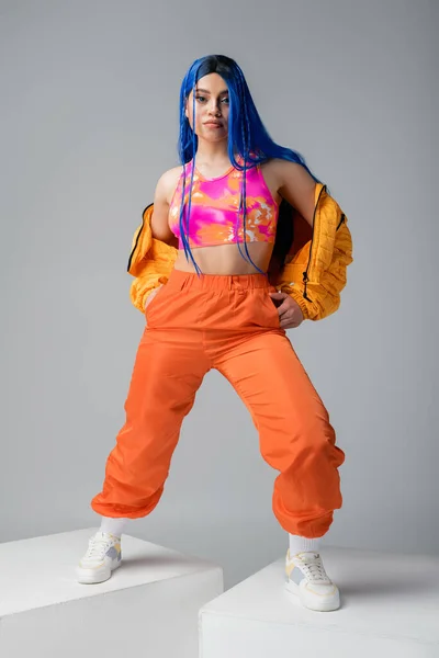Volle Länge, modisches Statement, Frau mit blauen Haaren posiert in Pufferjacke und orangefarbener Hose auf grauem Hintergrund, steht auf weißen Würfeln, leuchtende Farbe, weibliches Modell, urbane Mode — Stockfoto