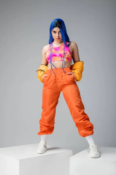 Full length, fashion forward, tattooed woman with blue hair posing with hands in pockets of orange pants on grey background, standing on white cubes, vibrant color, female model, urban fashion — Stock Photo