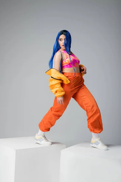 Повна довжина, мода вперед, молода жінка з блакитним волоссям позує в пуховику і помаранчеві штани на сірому фоні, ходьба на білих кубиках, яскравий колір, жіноча модель, міська мода — стокове фото