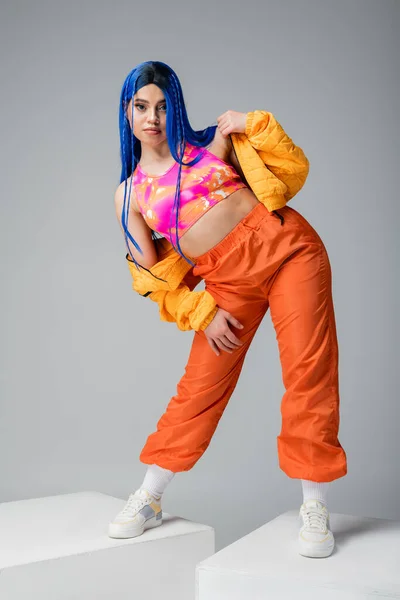 Comprimento total, moda para a frente, mulher com cabelo azul ajustando jaqueta puffer e de pé em calças laranja em cubos brancos fundo cinza, cor vibrante, modelo feminino, moda urbana — Fotografia de Stock