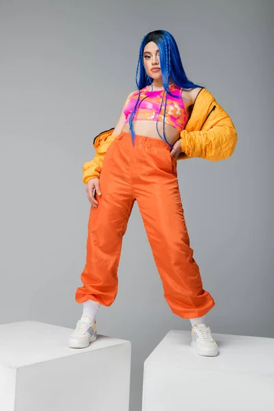 Volle Länge, Mode nach vorne, weibliches Model mit blauen Haaren posiert in Pufferjacke und orangefarbener Hose auf grauem Hintergrund, mit der Hand an der Hüfte auf weißen Würfeln stehend, lebendige Farbe, urbane Mode — Stockfoto