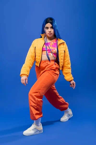 Volle Länge, Mode nach vorne, junges weibliches Model mit blauen Haaren posiert in Pufferjacke und orangefarbener Hose auf blauem Hintergrund, lebendige Farbe, urbane Mode, Individualismus — Stockfoto