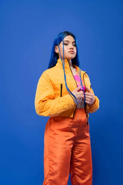 Cabelo tingido, moda para a frente, modelo feminino tatuado com cabelo azul posando em jaqueta puffer e calças laranja no fundo azul, cor vibrante, moda urbana, individualismo, jovem — Fotografia de Stock