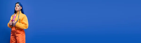 Capelli tinti, moda in avanti, modello femminile tatuato con capelli blu in posa in giacca di puffer e pantaloni arancioni su sfondo blu, colore vivace, moda urbana, individualismo, giovane donna, banner — Foto stock