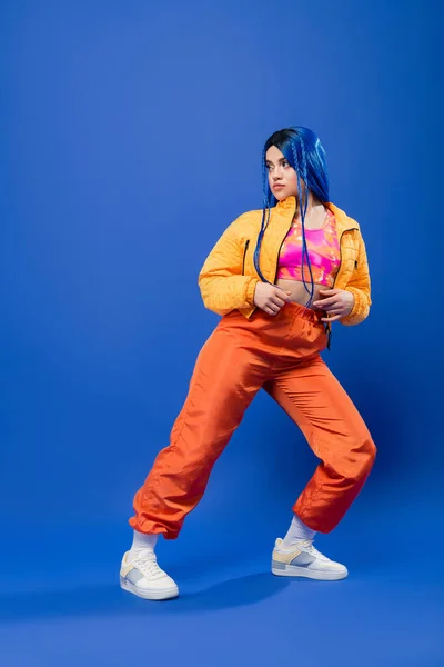 Comprimento total, cabelo tingido, moda para a frente, modelo feminino tatuado com cabelo azul posando na jaqueta puffer e calças laranja no fundo azul, cor vibrante, moda urbana, individualismo, jovem — Fotografia de Stock