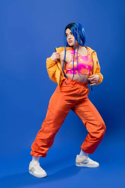 Volle Länge, gefärbte Haare, Mode und Stil, weibliches Model mit blauen Haaren posiert in Pufferjacke und orangefarbener Hose auf blauem Hintergrund, lebendige Farbe, urbane Mode, Individualismus, junge Frau — Stockfoto