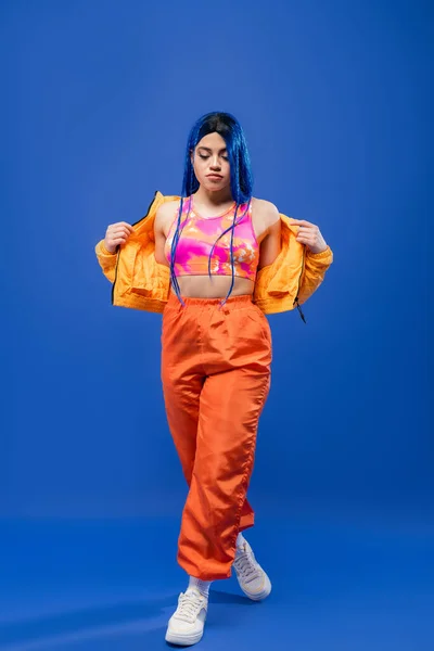 Full length, dyed hair, modern style, female model with blue hair posing in puffer jacket and orange pants on blue background, vibrant color, urban fashion, individualism, young woman — Stock Photo