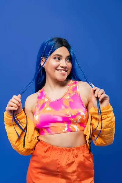 Beauty trends, dyed hair, cheerful female model with blue hair posing in puffer jacket on blue background, vibrant color, urban fashion, individualism, young woman smiling and looking away — Stock Photo