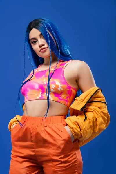 Colorful clothes, dyed hair, female model with blue hair posing with hands in pockets on blue background, puffer jacket, vibrant color, urban fashion, individualism, young woman with funky look — Stock Photo