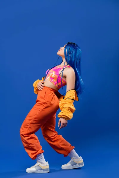 Volle Länge, Frau mit gefärbten Haaren, Mode nach vorne, weibliches Model mit blauen Haaren posiert in Pufferjacke und orangefarbener Hose auf blauem Hintergrund, lebendige Farbe, urbane Mode, Individualismus — Stockfoto