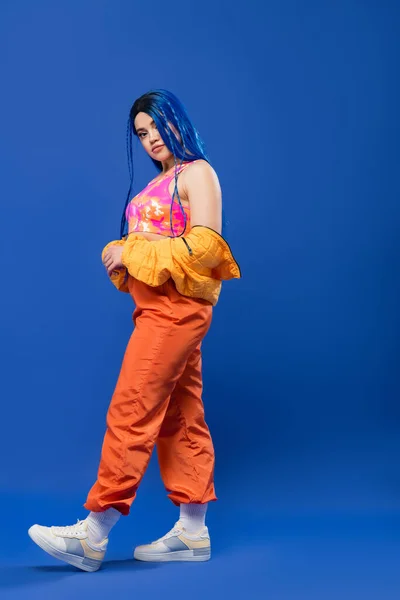 Longitud completa, mujer tatuada con el pelo teñido, la moda hacia adelante, modelo femenino con el pelo azul posando en chaqueta hinchable y pantalones de color naranja sobre fondo azul, color vibrante, moda urbana, individualismo - foto de stock