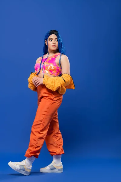 Volle Länge, Frau mit gefärbten Haaren, modisches Statement, tätowiertes weibliches Model mit blauen Haaren posiert in Pufferjacke und orangefarbener Hose auf blauem Hintergrund, lebendige Farbe, urbane Mode, Individualismus — Stockfoto