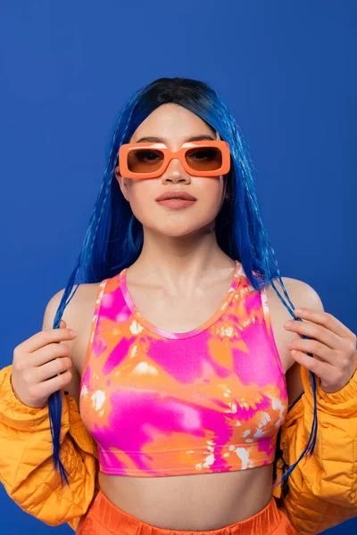 Mode-Statement, junges weibliches Model mit blauen Haaren und Zöpfen posiert mit trendiger Sonnenbrille auf blauem Hintergrund, Generation Z, Rebellentyp, bunte Kleidung, Individualismus, moderne Frau — Stockfoto