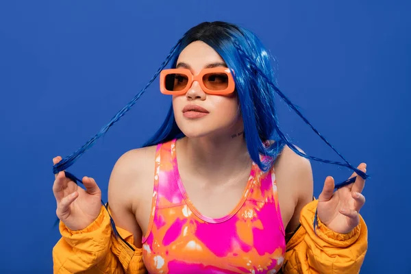 Fashion statement, young female model with blue hair touching braids and trendy sunglasses isolated on blue background, generation z, rebel style, colorful clothes, individualism, modern woman — Stock Photo