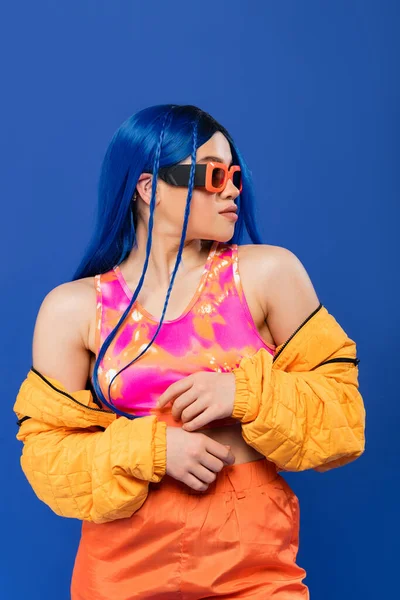 Fashion forward, young female model with blue hair and trendy sunglasses isolated on blue background, generation z, rebel style, colorful clothes, individualism, modern woman looking away — Stock Photo