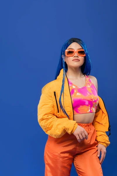 Gen z, fashion statement, young female model with blue hair adjusting trendy sunglasses isolated on blue background, rebel style, colorful clothes, individualism, modern woman — Stock Photo