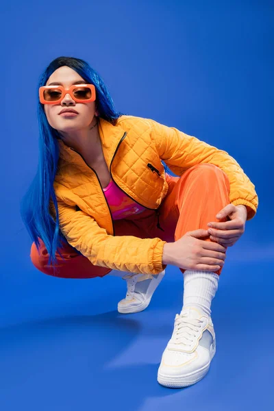 Full length of pretty young female model with blue hair and trendy sunglasses sitting on haunches on blue background, generation z, rebel style, individualism, modern fashion, trendy accessory — Stock Photo