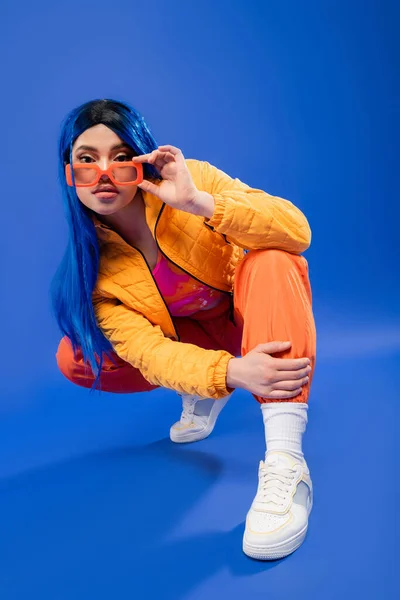 Individualism, full length of young female model with blue hair and trendy sunglasses sitting on haunches on blue background, generation z, rebel style, modern fashion, trendy accessory — Stock Photo