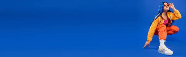 Generazione z, lunghezza intera di grazioso modello femminile con capelli blu e occhiali da sole alla moda seduti su fianchi su sfondo blu, stile ribelle, moda moderna, accessorio alla moda, banner — Foto stock