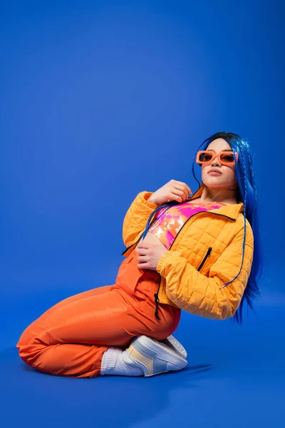 Declaração de moda de gen z, comprimento total de modelo feminino bonito com cabelo azul e óculos de sol na moda sentado no fundo azul, estilo rebelde, moda moderna, acessório da moda, juventude — Fotografia de Stock