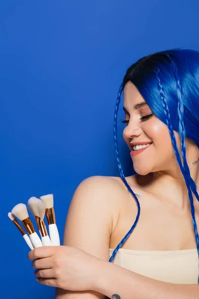 Beauty industry, individualism, joyful young woman with vibrant hair and eyes holding cosmetic brushes on blue background, makeup, beauty trends, visage, youth, self expression — Stock Photo