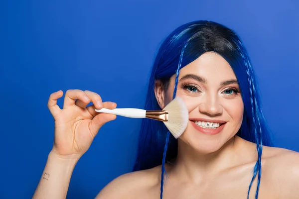 Beauty trends, individualism, cheerful young woman with vibrant eyes and hair looking at camera while holding makeup brush on blue background, cosmetic, self expression, visage, youth — Stock Photo