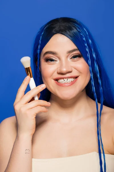 Individualism, joyous young woman with vibrant eyes and hair looking at camera while holding makeup brush on blue background, cosmetic, self expression, visage, youth, beauty trends — Stock Photo