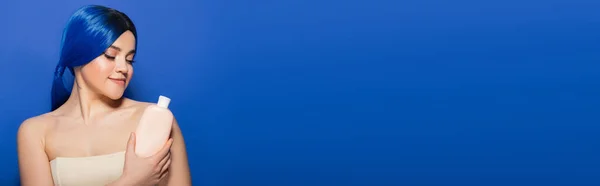 Körper- und Haarpflegekonzept, Porträt einer tätowierten jungen Frau mit lebendiger Haarfarbe, die mit nackten Schultern auf blauem Hintergrund posiert, Kosmetikflasche mit Shampoo in der Hand, Beauty-Trends, Banner — Stockfoto