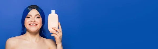 Schönheitstrends, Haarpflegekonzept, Porträt einer glücklichen jungen Frau mit lebendiger Haarfarbe, die mit nackten Schultern auf blauem Hintergrund posiert, Kosmetikflasche mit Shampoo in der Hand, Werbung, Banner — Stockfoto