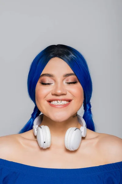 Amante de la música, mujer joven positiva con el pelo azul y auriculares inalámbricos sonriendo sobre fondo gris, juventud vibrante, individualismo, subcultura moderna, expresión personal, tatuaje, sonido - foto de stock