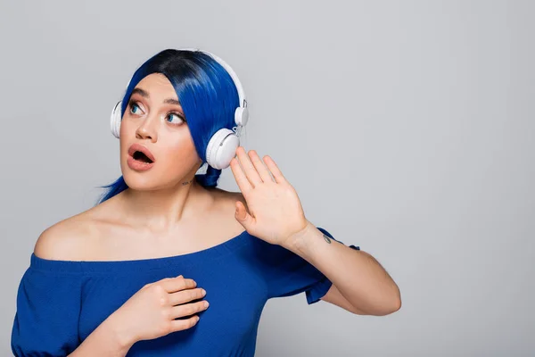 Expresión personal, mujer joven conmocionada con el pelo azul escuchando música en auriculares inalámbricos sobre fondo gris, juventud vibrante, individualismo, subcultura moderna, tatuaje, sonido - foto de stock