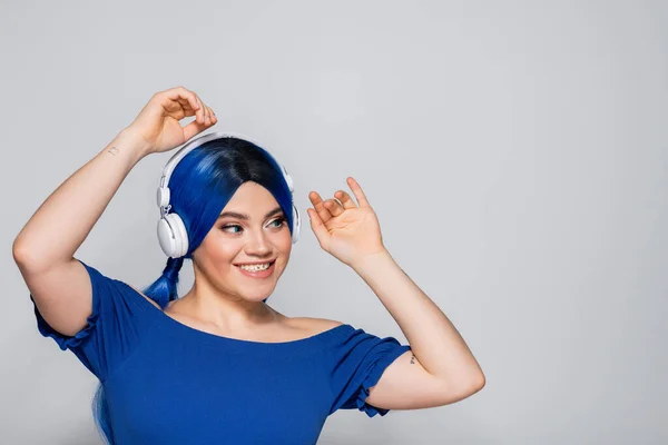 Expresión personal, mujer joven positiva con cabello azul escuchando música en auriculares inalámbricos sobre fondo gris, juventud vibrante, individualismo, subcultura moderna, tatuaje, sonido - foto de stock
