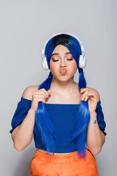 Самовираження, молода жінка з блакитним волоссям слухає музику в бездротових навушниках на сірому фоні, гладить губи, яскрава молодь, індивідуалізм, сучасна субкультура, татуювання, звук — стокове фото