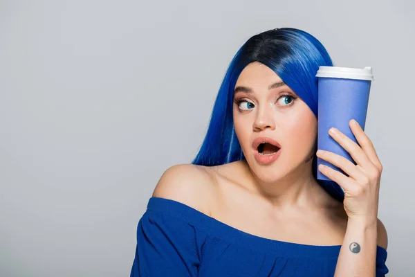 Para llevar taza, mujer joven asombrada con cabello azul y ojos sosteniendo café para ir sobre fondo gris, comida para llevar, cafeína, energía, tatuaje, color vibrante, expresión personal, individualismo - foto de stock