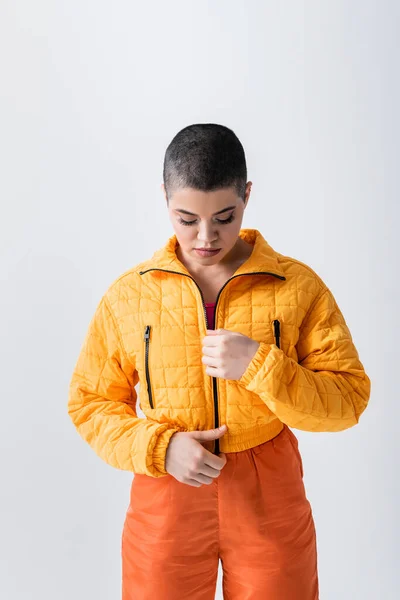 Outerwear, young woman with short hair zipping yellow puffer jacket on grey background, urban fashion, vibrant youth, trendy outfit, stylish look, studio photography, modern subculture — Stock Photo