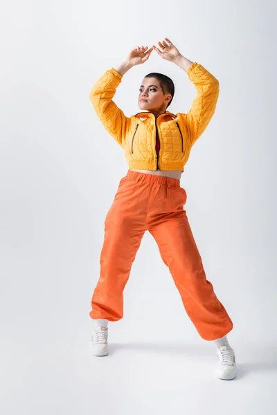 Outerwear, casual attire, puffer jacket and trendy orange pants, young tattooed model posing with raised hands on grey background, woman with short hair, generation z — Stock Photo