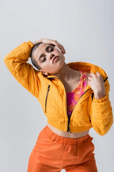 Outerwear, individualism, fashion model posing with closed eyes, young woman with short hair standing in yellow puffer jacket on grey background, isolated, youth culture, casual wear — Stock Photo