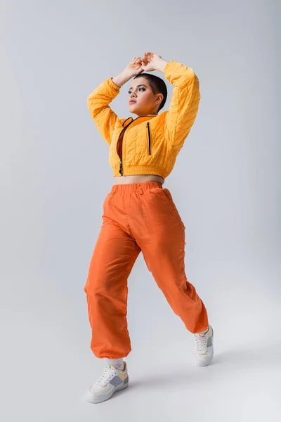 Modern subculture, outerwear, casual attire, fashion model posing in yellow puffer jacket and orange pants on grey background, woman with short hair with raised hands, stylish look, individualism — Stock Photo