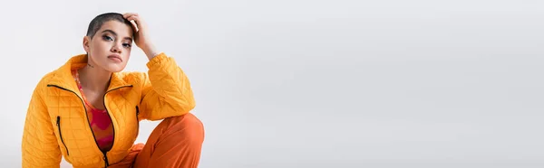 Urbane Mode, Oberbekleidung, weibliches Model posiert in Freizeitkleidung, junge Frau mit kurzen Haaren und Pufferjacke sitzt auf grauem Hintergrund, blickt in die Kamera, persönlicher Stil, tätowiert, Banner — Stockfoto