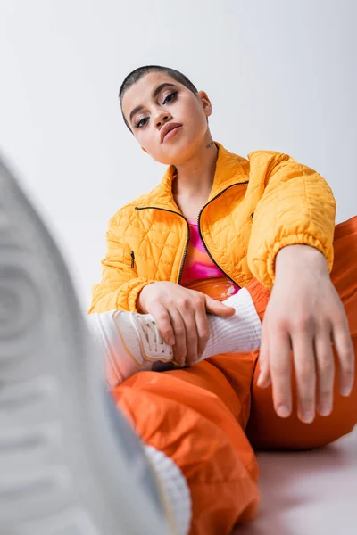Outerwear, declaração de moda, tatuado jovem mulher em roupas coloridas sentado e olhando para a câmera em fundo cinza, estilo urbano, individualismo, energia vibrante e jovem, foreground borrado — Fotografia de Stock