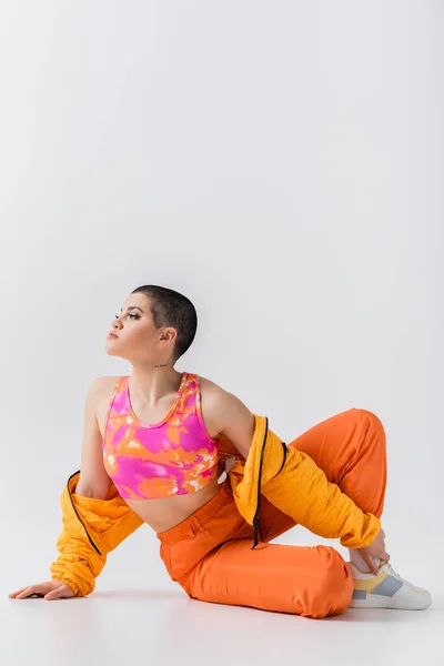 Vibrant youth, tattooed young woman with short hair sitting on grey background, generation z, fashion forward, colorful clothes, female model with personal style, self expression, trendy outfit — Stock Photo