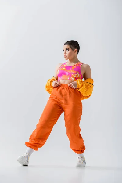 Female model, warm clothing, fashionable young woman with short hair posing in puffer jacket and vibrant pants on grey background, outerwear, modern subculture, youthful energy, full length — Fotografia de Stock