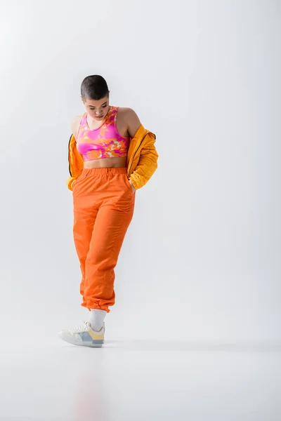 Современная субкультура, модная молодая женщина с короткой парой позирующих рук в карманах оранжевых брюк на сером фоне, пиджак фугу. верхняя одежда, юная энергия, полная длина — стоковое фото