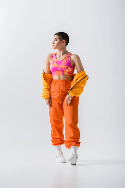 Generation z, fashionable young woman with short hair posing in puffer jacket and vibrant pants on grey background, looking away, outerwear, modern subculture, youthful energy, full length - foto de stock