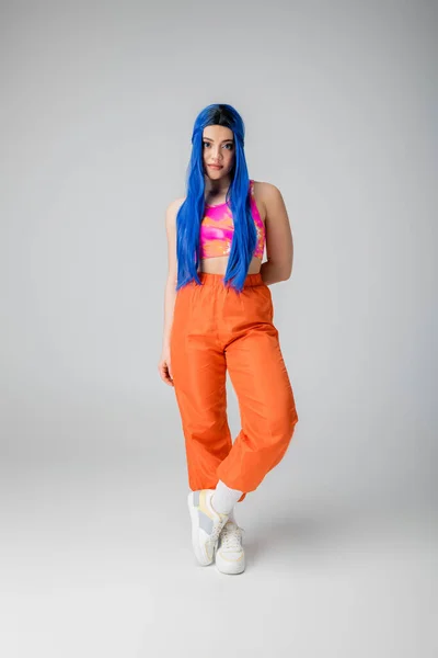 Повна довжина молодої жінки з блакитним кольором волосся позує в стильному вбранні на сірому фоні, довге волосся, помаранчеві штани та рожевий корінь зверху, мода вперед, особистий стиль, індивідуалізм — стокове фото