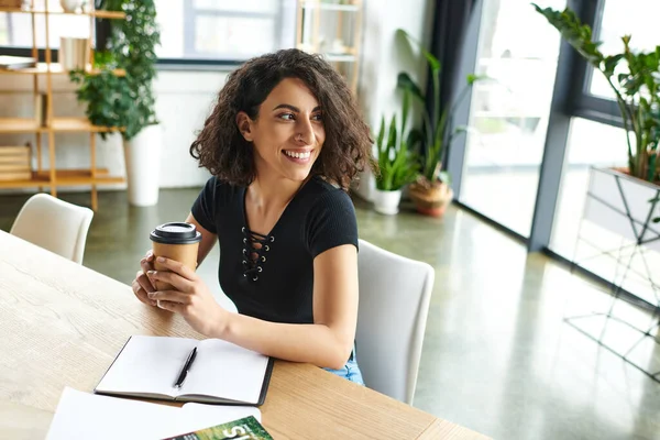 Cheerful multiracial couch holding coffee to go and looking away while sitting at table with notebook and pen in cozy environment of women interest club, social activism and dedication concept — Stock Photo