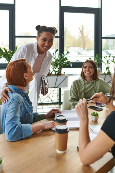 Joven afroamericano motivación entrenador con portátil sonriendo cerca diverso grupo de miembros multiétnicos de club de interés de las mujeres cerca de café en tazas de papel, apoyo mutuo y concepto de comprensión - foto de stock