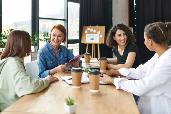 Positive redhead woman holding magazine during communication with multiethnic female friends at table with coffee to go and notebooks in women interest club, leisure and knowledge-sharing concept — Stock Photo