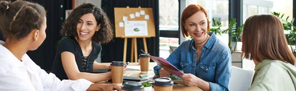 Happy redhead woman sitting with magazine near table with takeaway drinks and group of multiethnic girlfriends talking in interest club, positive communication and knowledge-sharing concept, banner — Stock Photo